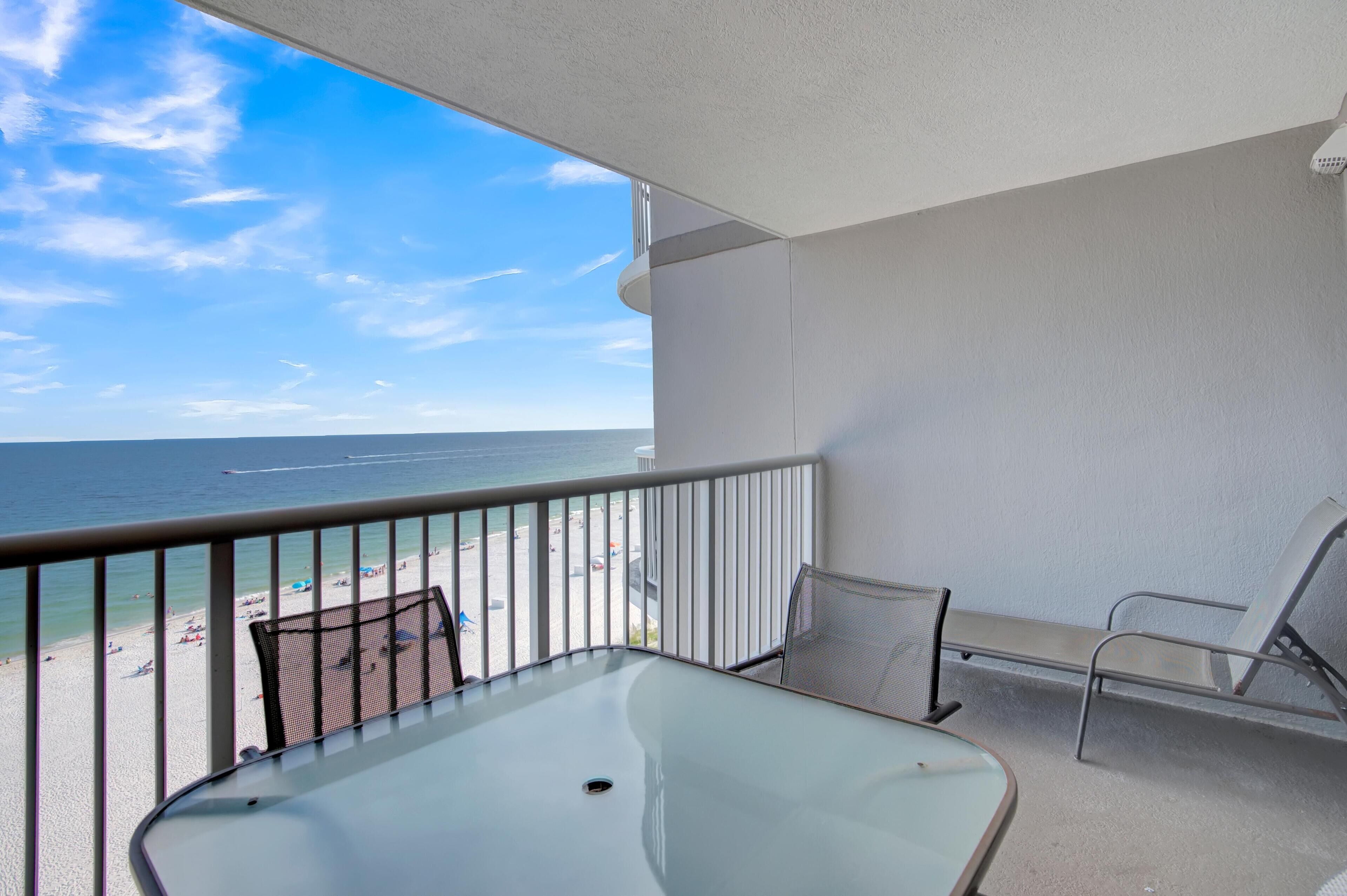 A balcony with a glass table and two chairs, overlooking a beach with people and a clear sky.