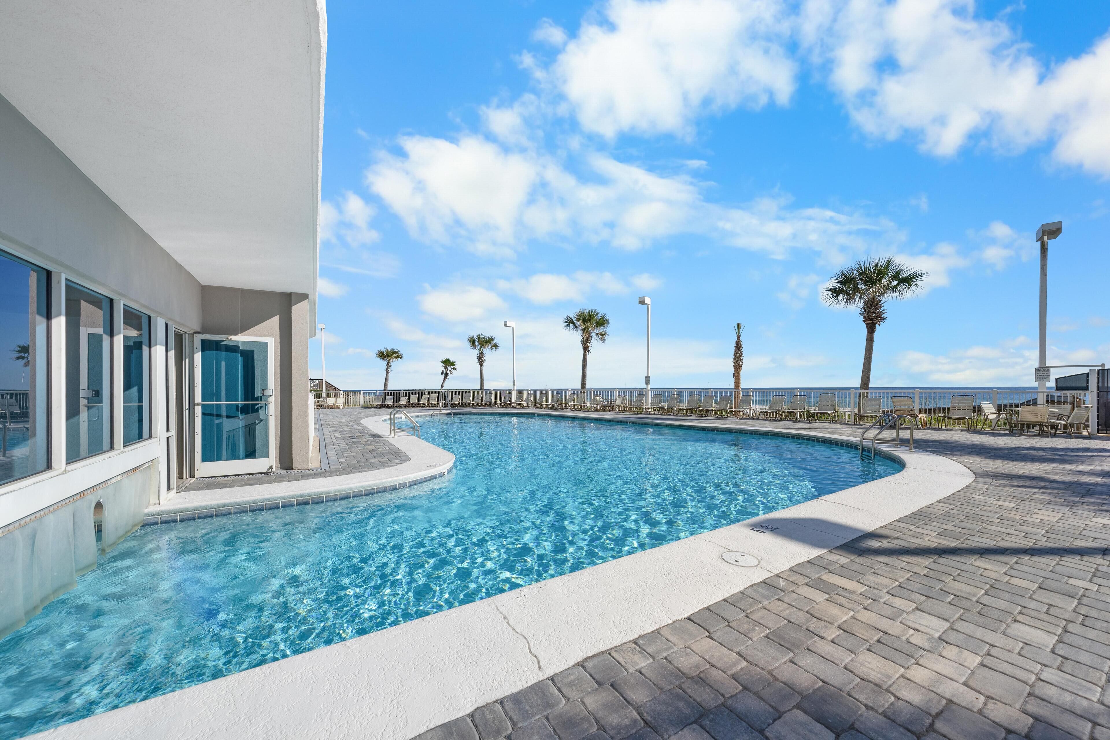 A swimming pool with clear blue water, surrounded by a paved deck and palm trees.