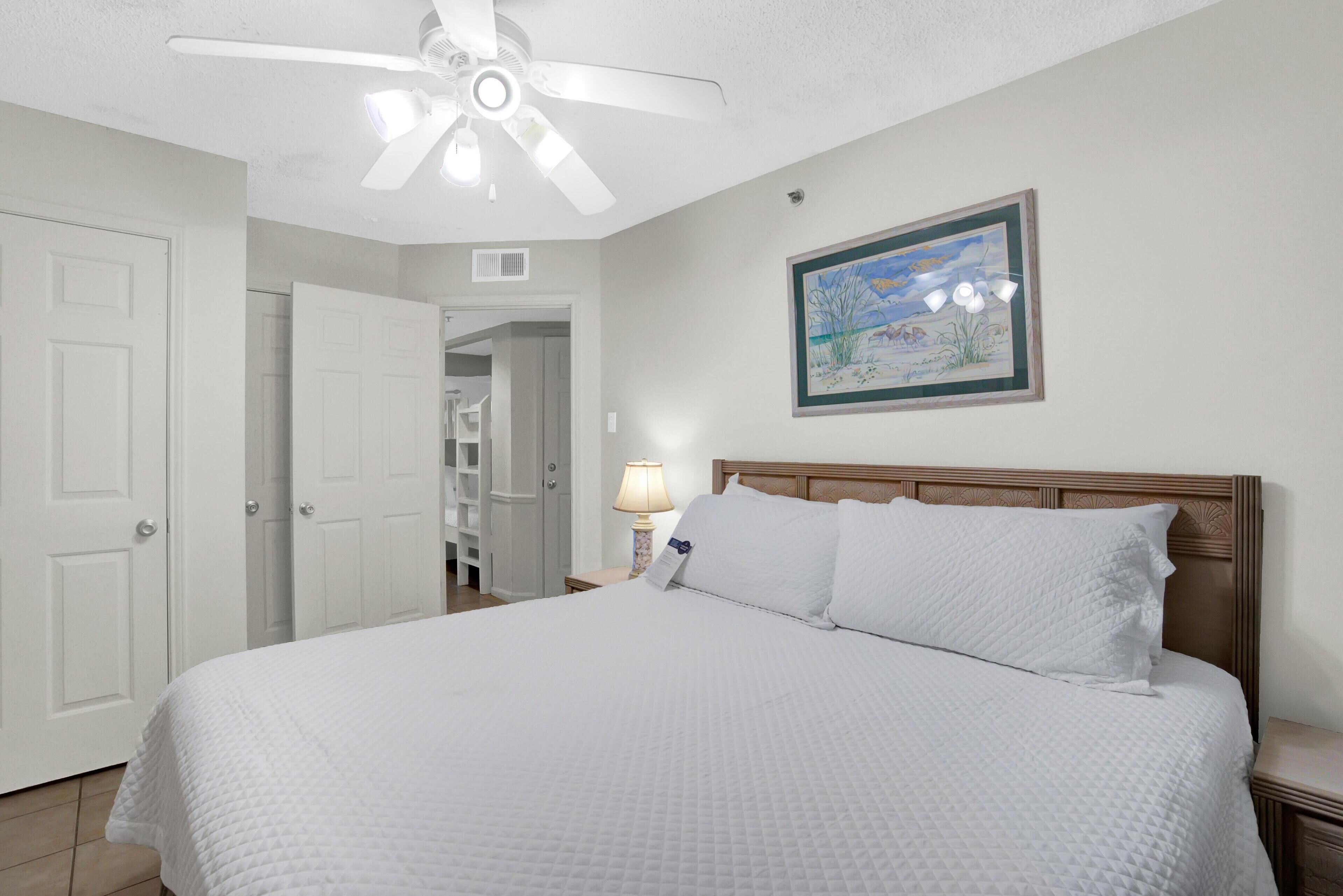 A bedroom with a large bed, a ceiling fan, a nightstand, a lamp, and a framed picture on the wall.