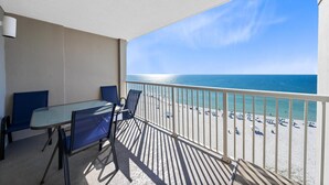 Ein Balkon mit Glastisch und Stühlen, der Blick auf einen Strand mit Sonnenschirmen und einem klaren Himmel bietet.