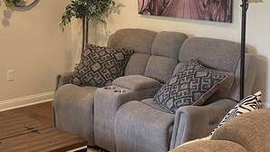 Ein Wohnzimmer mit einem grauen Ecksofa, einem Holztisch, einer Stehlampe, einem Wandbild mit Weinfässern und einem Topfpflanzen.