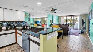A kitchen with a central island, a dining area with a sofa and chairs, and a view of the outdoors through large windows. - Wind Drift 114n (Orange Beach)