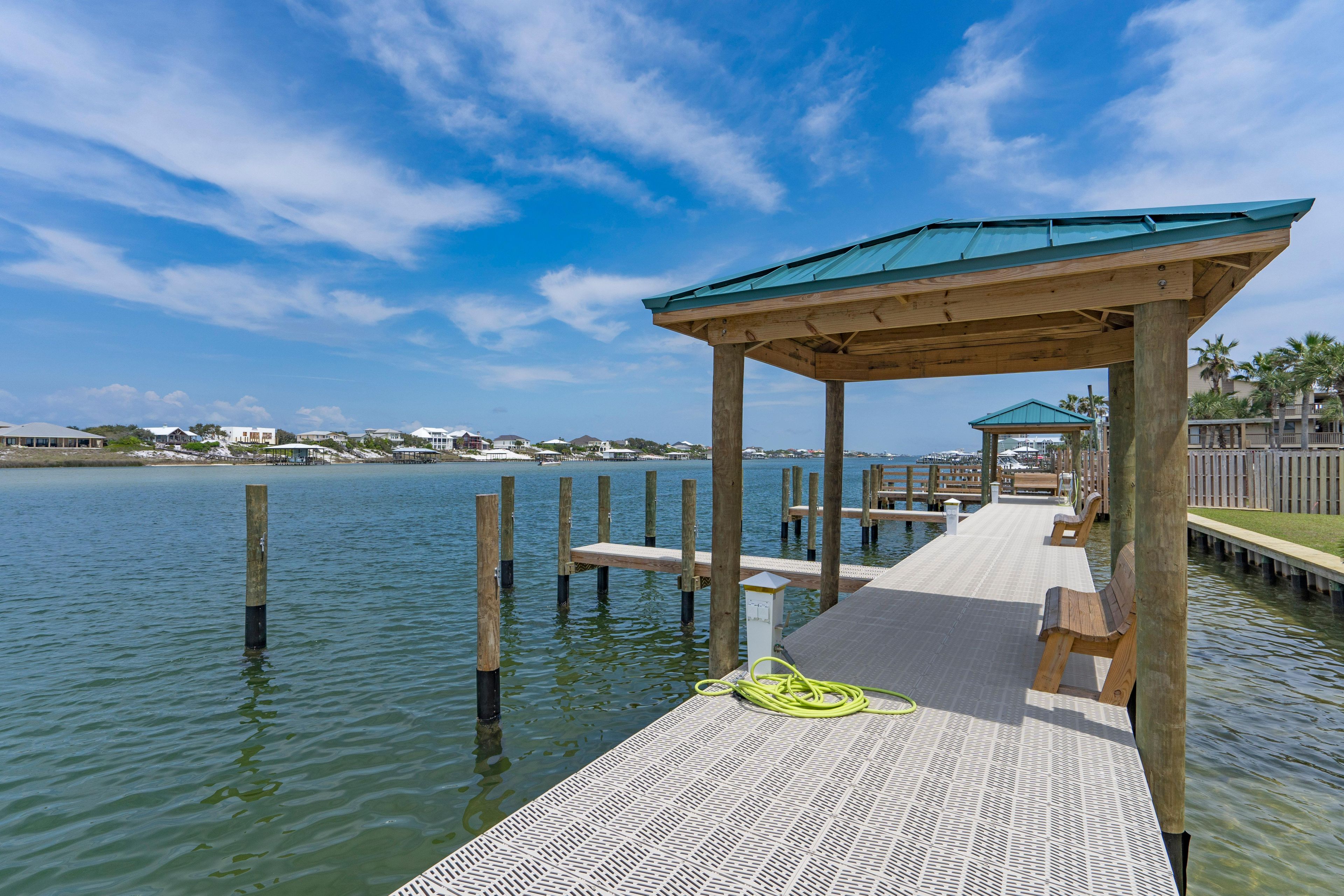 A wooden pier with a covered seating area, extending into calm water, with a clear sky above.