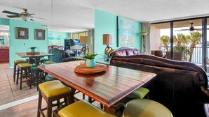 A dining area with a wooden table, green and yellow chairs, and a black leather booth. There are ceiling fans, a large window with a view of palm trees, and artwork on the walls. - Wind Drift 114n (Orange Beach)
