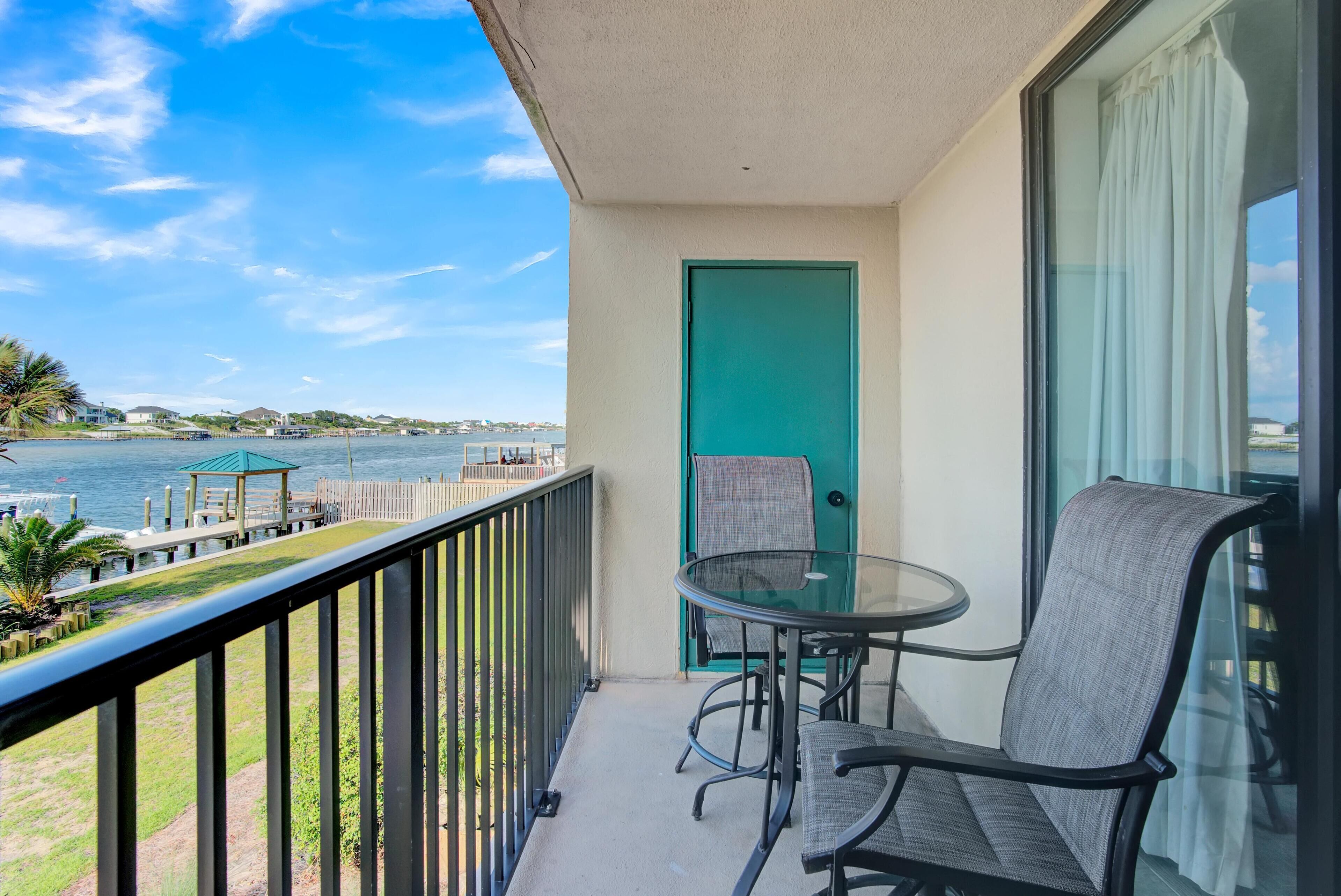 A balcony with a glass table and chairs, a teal door, and a view of the water and distant buildings.