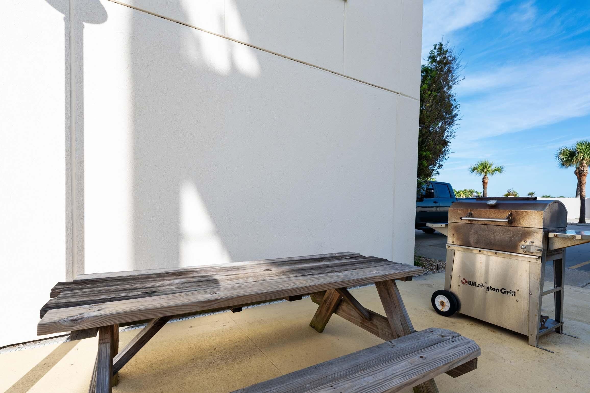 A wooden picnic table with a bench, a barbecue grill, and a vehicle in the background.