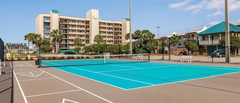 A tennis court with a net, surrounded by a fence, and a building with multiple balconies in the background.