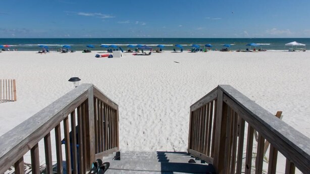 A beach with a wooden walkway leading to the sand. Beach umbrellas and chairs are visible in the distance. - Wind Drift 114n (Orange Beach)