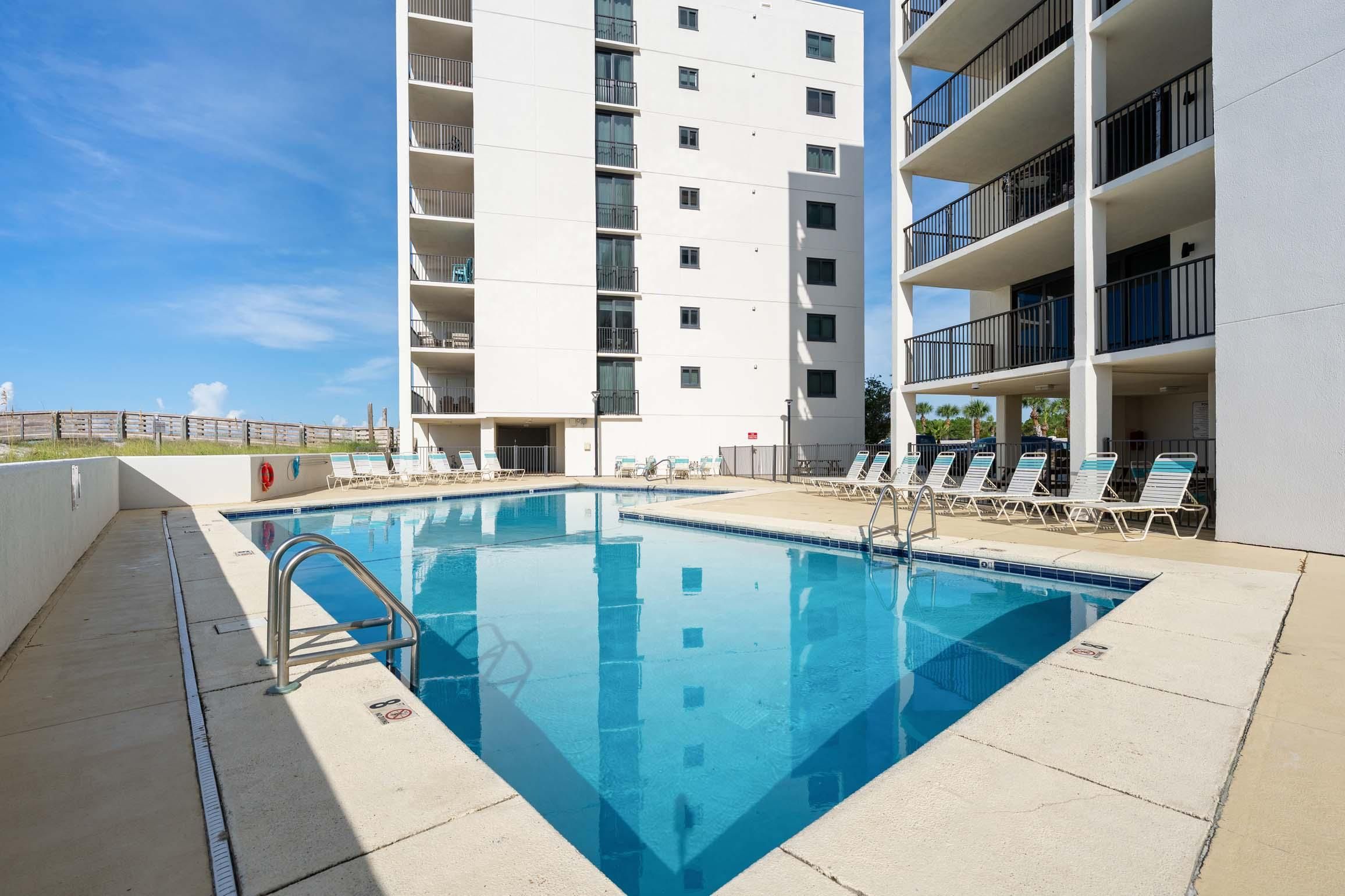 A swimming pool with a metal ladder, surrounded by lounge chairs and a multi-story building with balconies.
