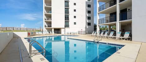 A swimming pool with a metal ladder, surrounded by lounge chairs and a multi-story building with balconies.