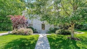Una casa de piedra con un sendero de piedra que lleva a la entrada, rodeada de céspedes verdes y diversos arbustos.
