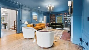 Un salón con una pared azul, un sofá amarillo, un sofá verde y un sillón blanco. Hay estanterías llenas de libros y una araña.