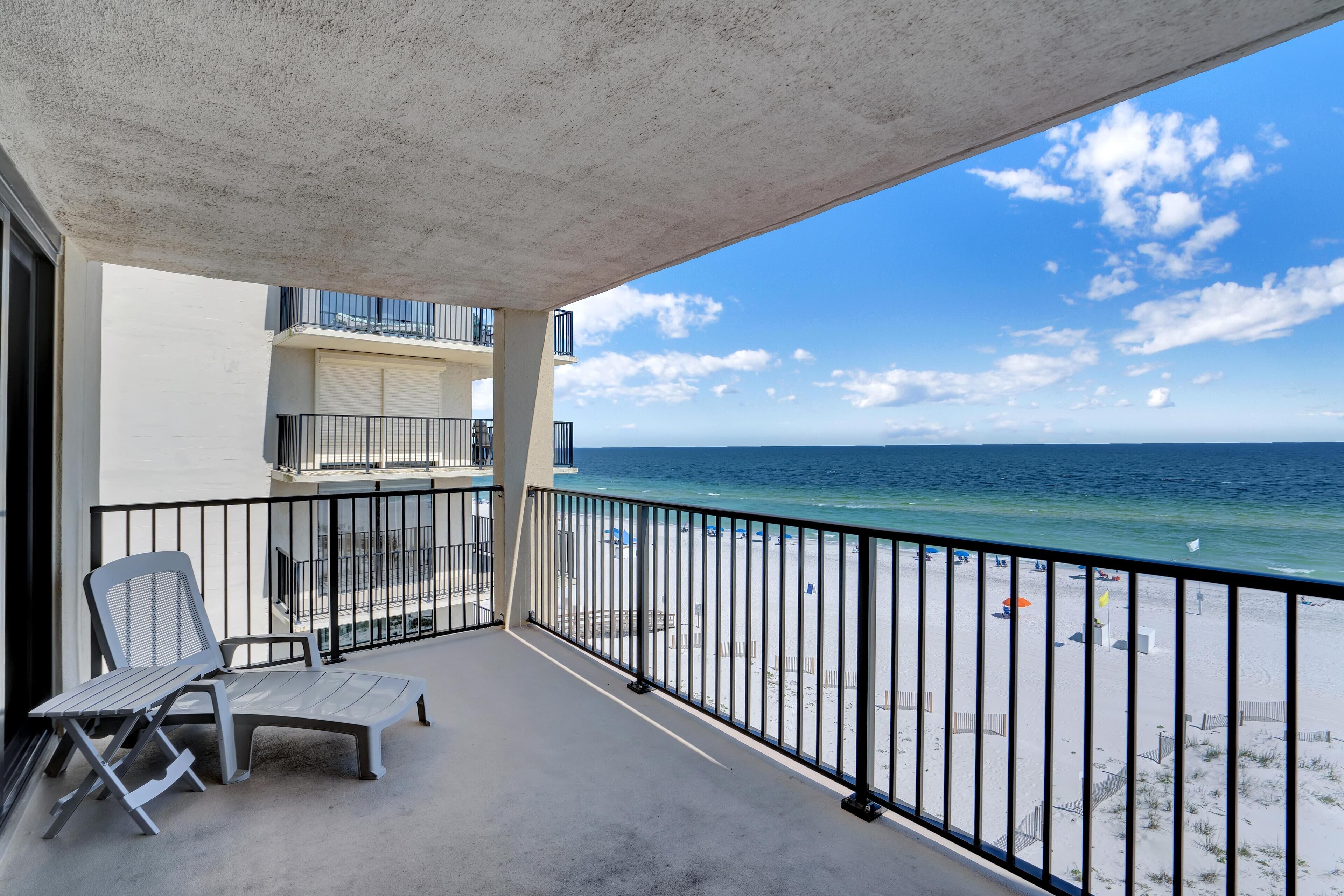A balcony with a view of the beach, a chair, and a table.