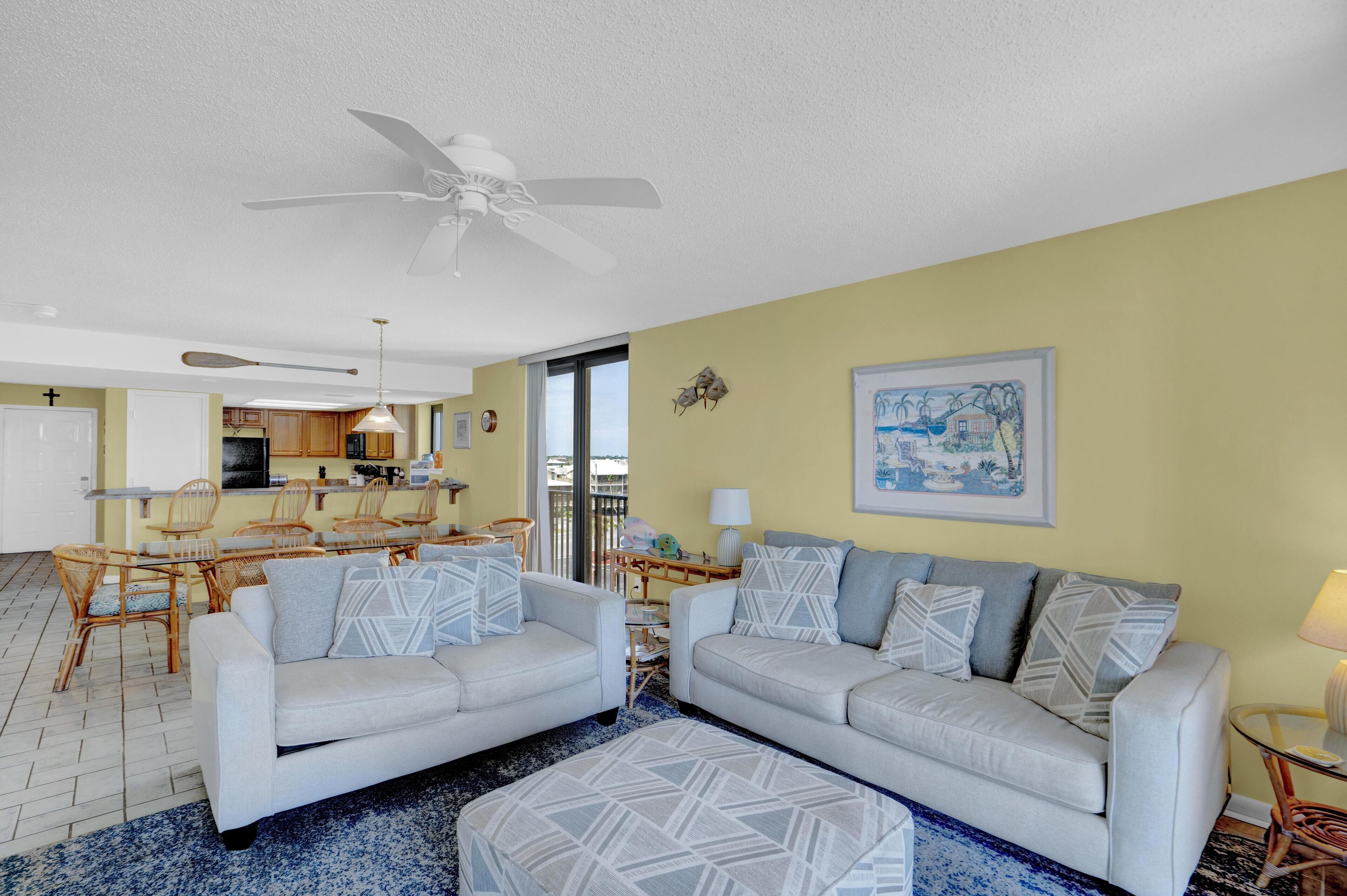 A living room with two sofas, a dining area with a table and chairs, a kitchen, and a ceiling fan.