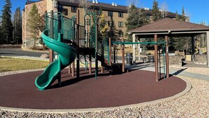 Un patio de juegos con un tobogán verde y estructuras para escalar, rodeado de una zona pavimentada y un edificio al fondo.