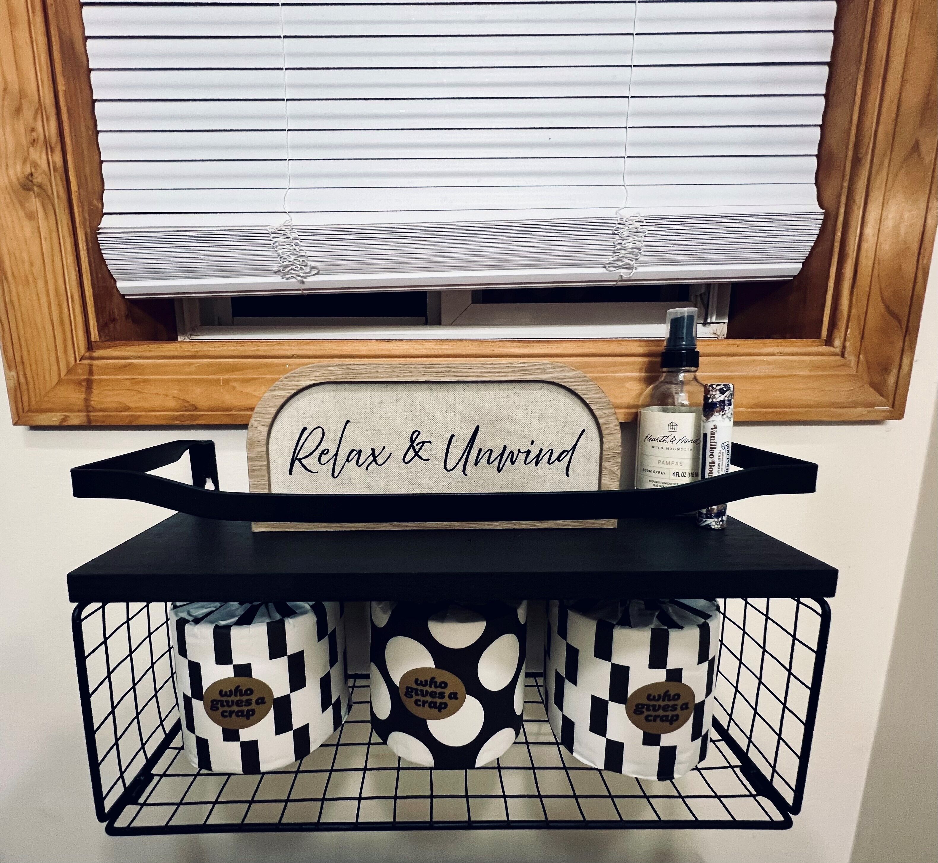 A wooden shelf with a sign saying "Relax & Unwind" above a window with blinds. Below, there are two storage bins with a checkered pattern and a bottle of hand sanitizer.