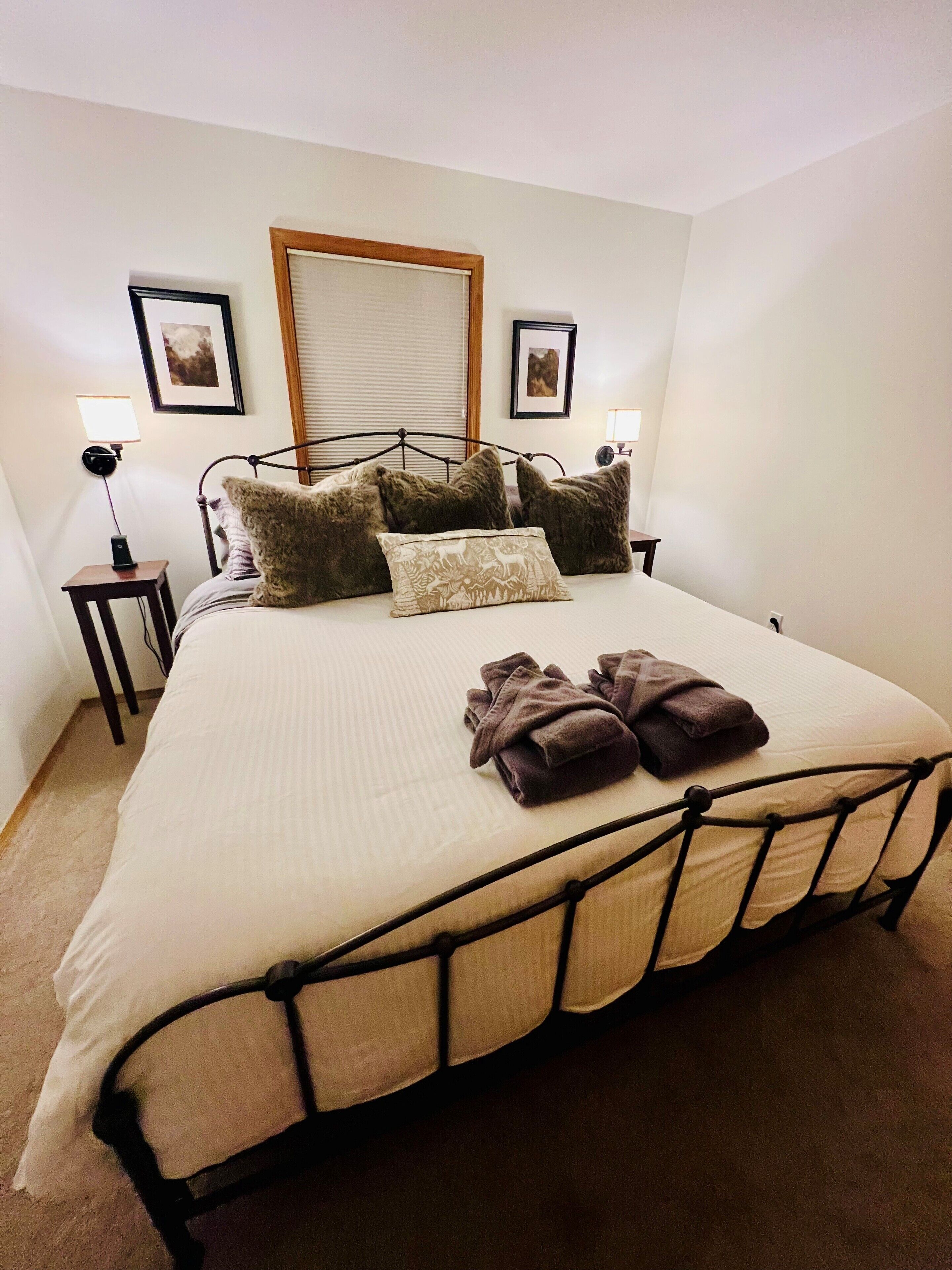 A neatly made bed with a metal frame, multiple pillows, and a blanket. There are two bedside tables with lamps, and a framed picture on the wall.