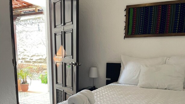 Habitación de hotel con cama, mesita de noche, lámpara, un cuadro en la pared y una puerta abierta que da a un espacio exterior con plantas.