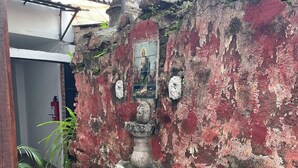 Un patio con una fuente de piedra, un muro de ladrillo rojo con un cuadro religioso y un suelo de baldosas.