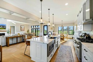 Une cuisine moderne dotée d’un îlot central, d’appareils électroménagers en acier inoxydable, de luminaires suspendus et de grandes fenêtres.