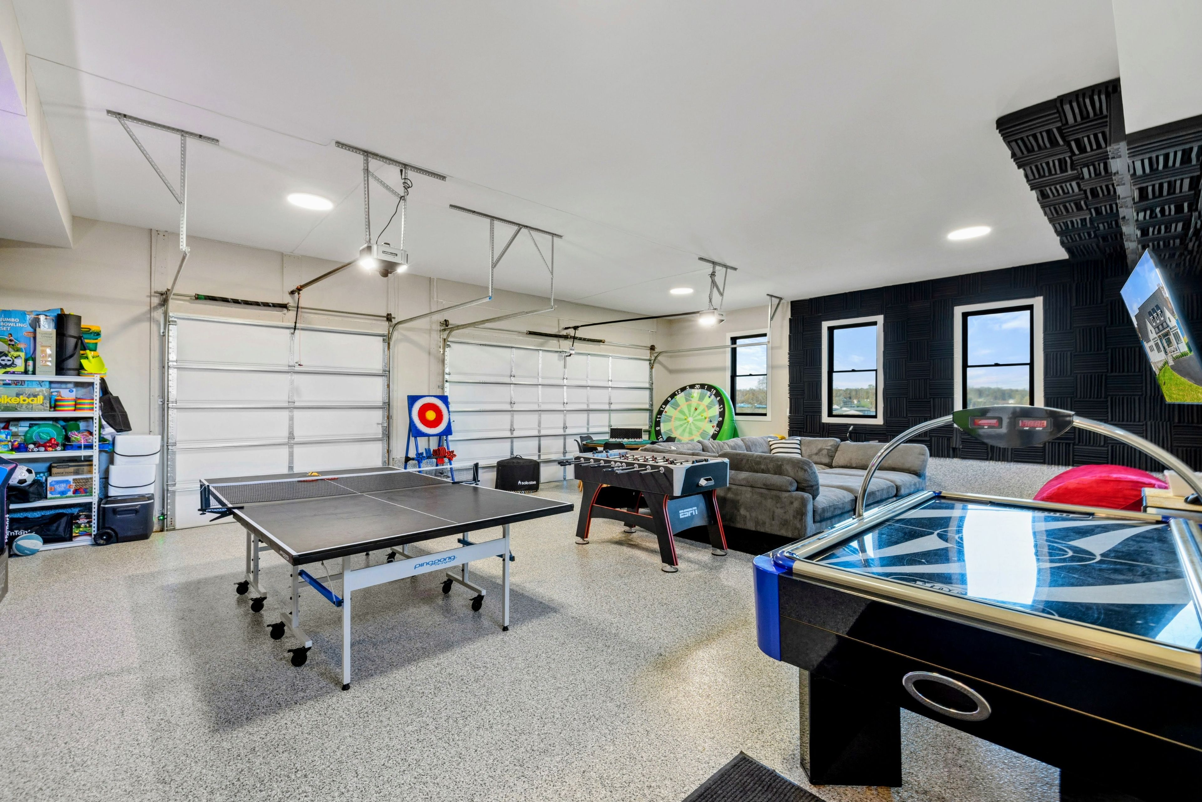 A recreational room with a ping pong table, foosball table, dartboard, and shelves with sports equipment.