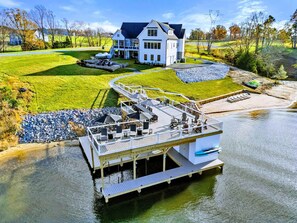 Une propriété au bord du lac comprenant une grande maison, un quai et une terrasse avec des meubles d’extérieur.