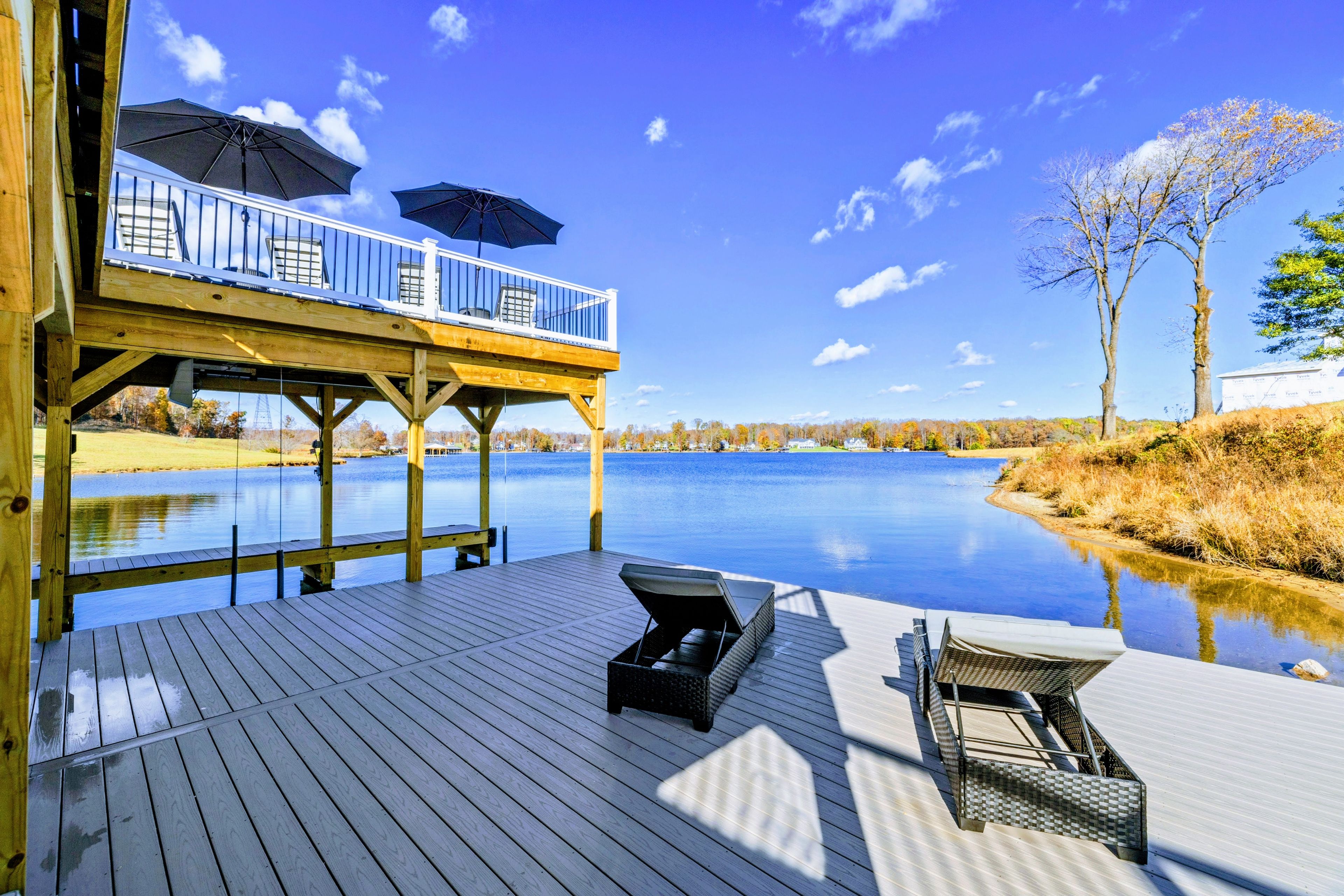A wooden deck with a view of a calm lake, a covered seating area with umbrellas, and a lounge chair.