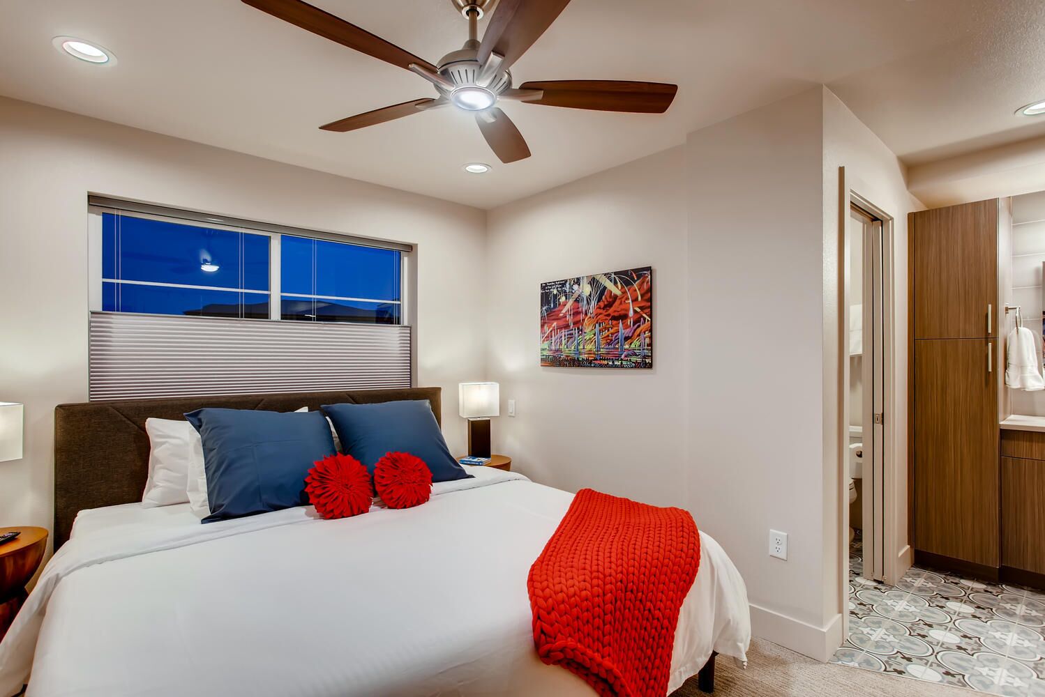 A bedroom with a bed, a ceiling fan, a window with blinds, a painting, and a wardrobe.