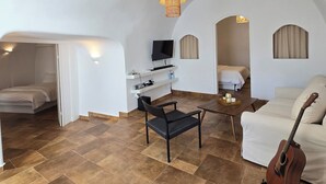 A spacious living area with a high ceiling, a dining table, a sofa, a guitar, and a television. - Charming 3-bedroom cave house in enchanting Fira with AC, WiFi, Washer/Dryer (Karterádos)