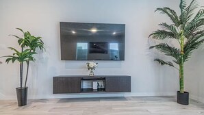A modern living room with a flat-screen TV mounted on the wall, a dark wooden console table with a vase of flowers, and two large potted plants. - Charming 3-Bedroom Luxury Home 5Min -> Downtown, NRG and Medical Center. (Houston)