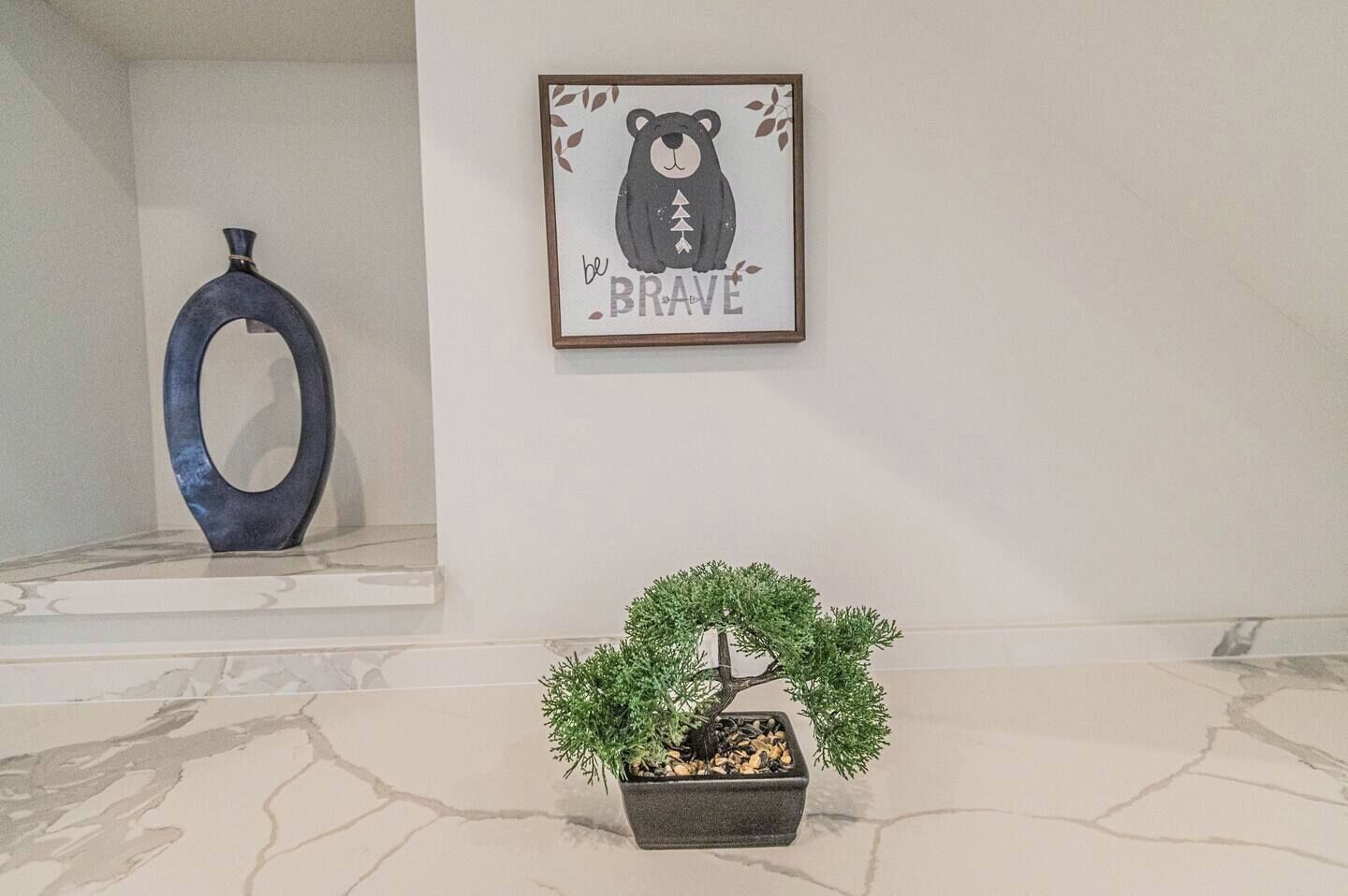 A room with a framed bear illustration on the wall, a decorative vase, and a bonsai tree on a tiled floor.