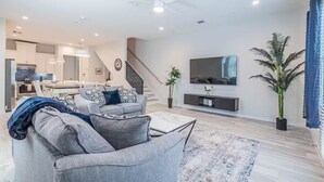 A modern living room with a grey sofa, a coffee table, and a TV. There's a staircase leading to the kitchen, and a large potted plant in the corner. - Charming 3-Bedroom Luxury Home 5Min -> Downtown, NRG and Medical Center. (Houston)