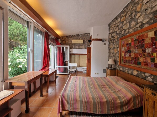 A bedroom with a bed, a desk, and a chair. There is a window with curtains, a stone wall, and a quilt on the wall.