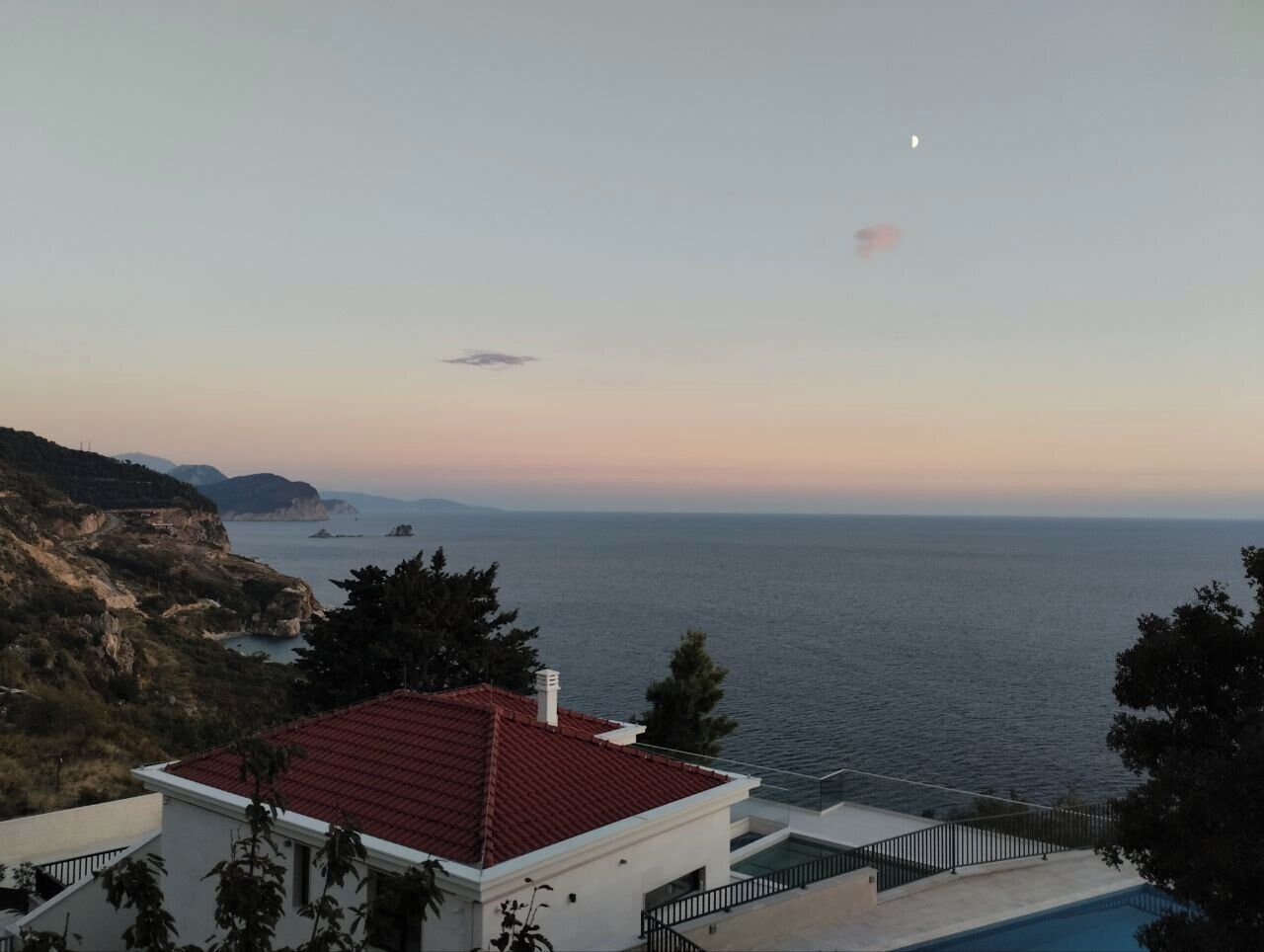 Vista costeira com um edifício de telhado vermelho, piscina e mar aberto com ilhas ao longe.