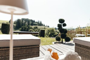 Eine Holzterrasse mit Korbmöbeln und einer Lampe, mit Blick auf einen angelegten Garten mit Holzzaun und Bäumen.