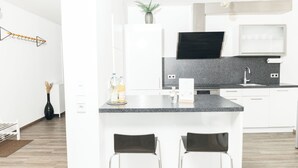 Et moderne køkken med hvide skabe, en granitbordplade og en backsplash i sort farve. Der er to barstole, en plante på bordpladen og en hængende lampe.