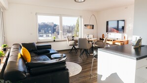 En moderne stue med en sort lædersofa, et spiseområde med bord og stole, et køkken med en bordplade og et fjernsyn monteret på væggen.