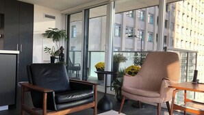 A modern living room with a black leather sofa, a woven armchair, and a small wooden table. There are potted plants and a view of a building through large windows. - Beautiful Light-filled Apartment with Spectacular Views of Toronto (Toronto)