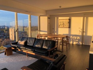 Ein modernes Wohnzimmer mit einem schwarzen Ledersofa, einem Holztisch und einem Esstisch mit Stühlen. Große Fenster bieten Blick auf die Stadt.