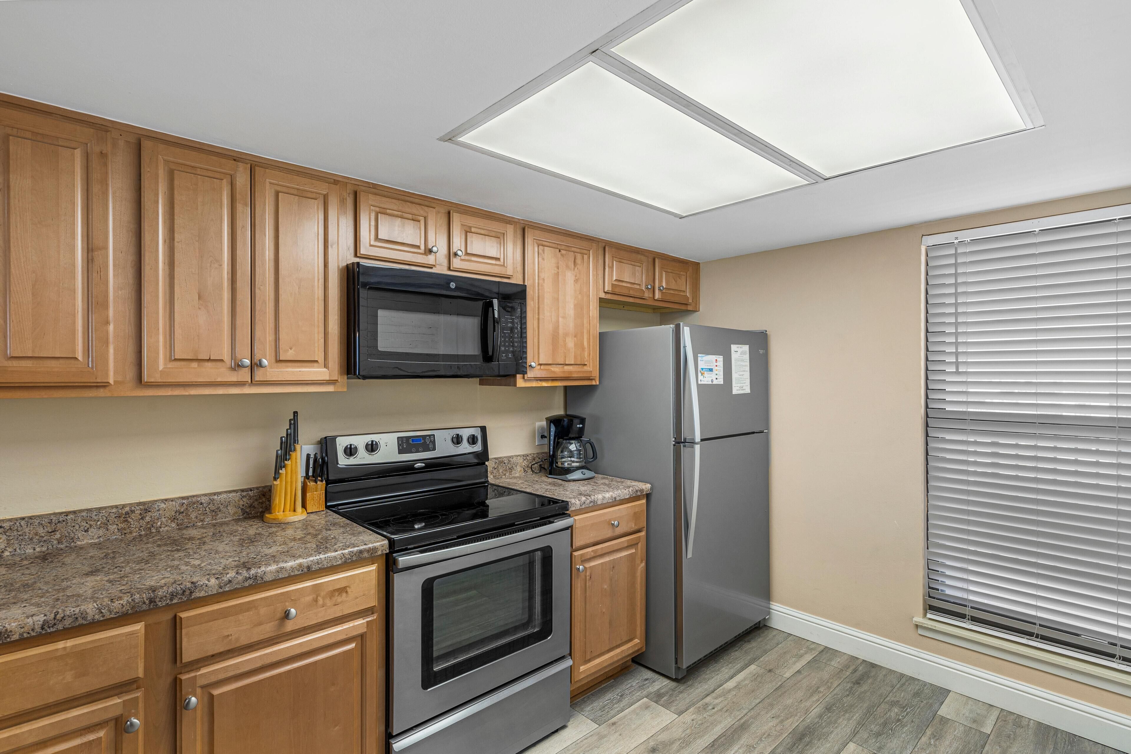 A kitchen with wooden cabinets, a black microwave, a stainless steel refrigerator, and a granite countertop.