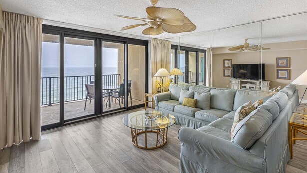 Un salon spacieux avec un grand canapé, une table basse en verre et un ventilateur de plafond. Il y a un balcon avec vue sur l’océan.