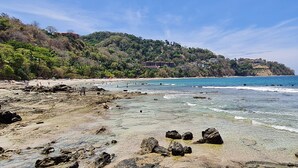 Ein Strand mit klarem Wasser, felsigem Ufer und Gebäuden an einem Hügelhang.