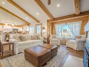 A spacious living room with a large sofa, two armchairs, and a brown leather ottoman. The room features wooden beams, a kitchen in the background, and large windows with sheer curtains. - The Cabin: Private Pool, Luxury Log Home (Grand Junction)