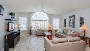 A spacious living room with a ceiling fan, large windows, a television, and a fireplace. - Villas at Island Club Community, pools, jacuzzi and more! (Kissimmee)