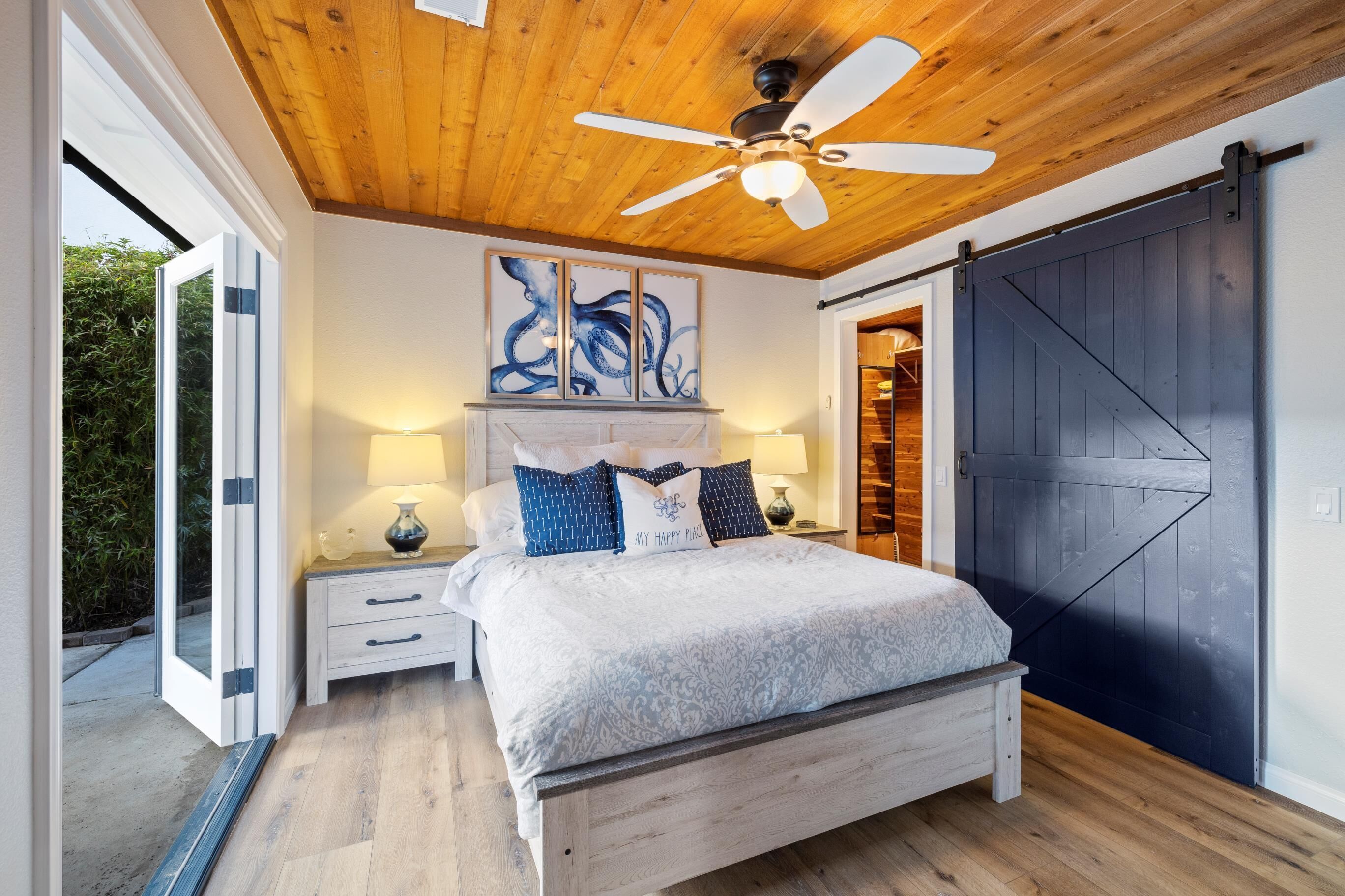 A bedroom with a wooden ceiling, a bed with a white and blue patterned bedspread, two bedside lamps, and a large artwork above the bed.
