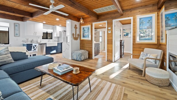 A spacious living area with a wooden ceiling, a sofa, a coffee table, and a dining table. There is a kitchen in the background with white cabinets and a refrigerator. - Cottage by the Harbor (Dana Point)