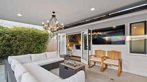 A modern outdoor living area with a white sectional sofa, two wooden chairs, a glass-top coffee table, and a hanging light fixture. - Cottage by the Harbor (Dana Point)