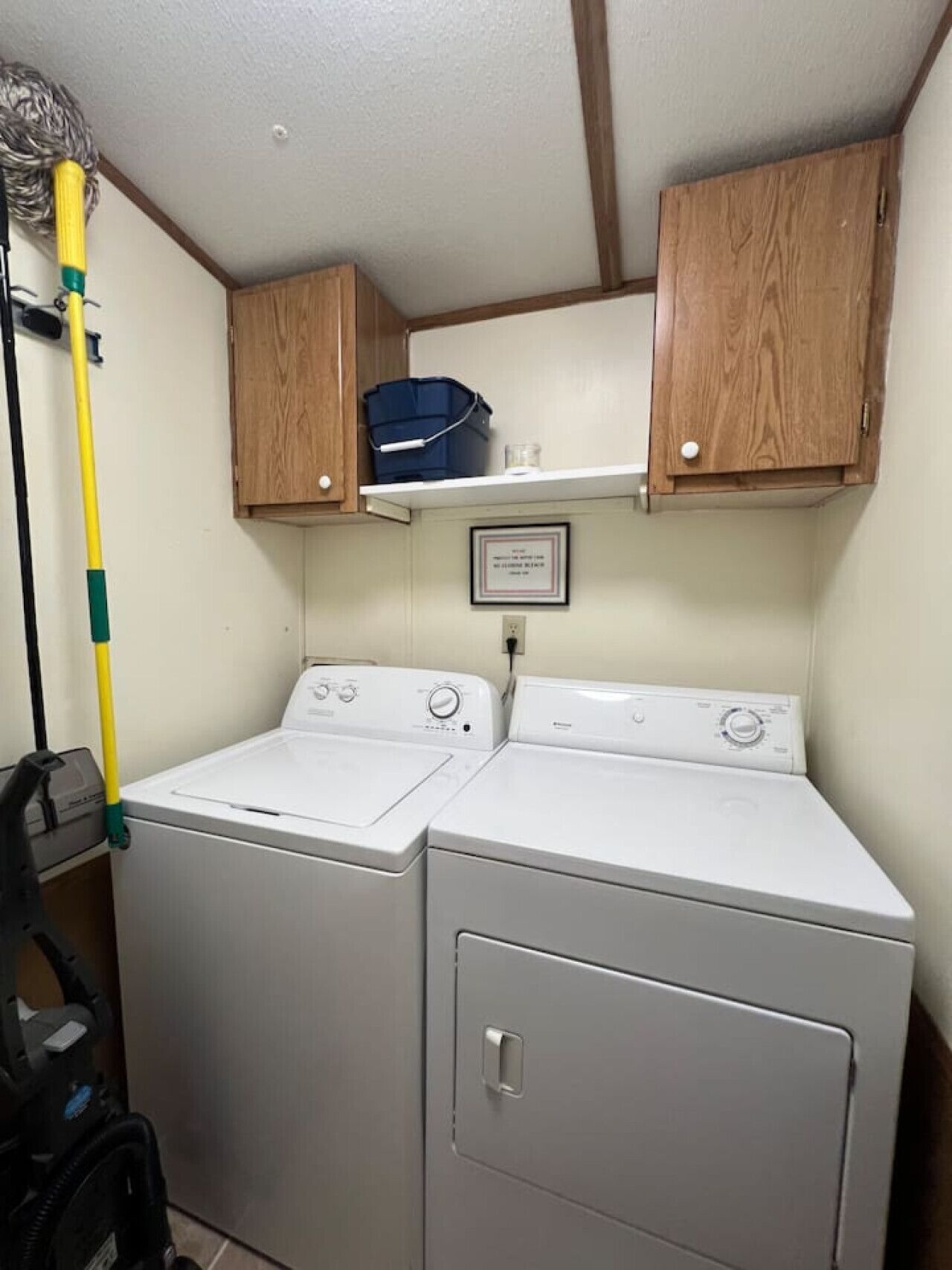 A laundry area with two washing machines, wooden cabinets above, and a mop on the left.
