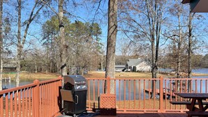 A wooden deck with a grill and picnic table, overlooking a lake with trees and a house in the background.