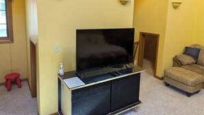 Una sala de estar con una televisión de pantalla plana sobre un soporte negro, un taburete rojo, un sofá color beige y una puerta de madera.